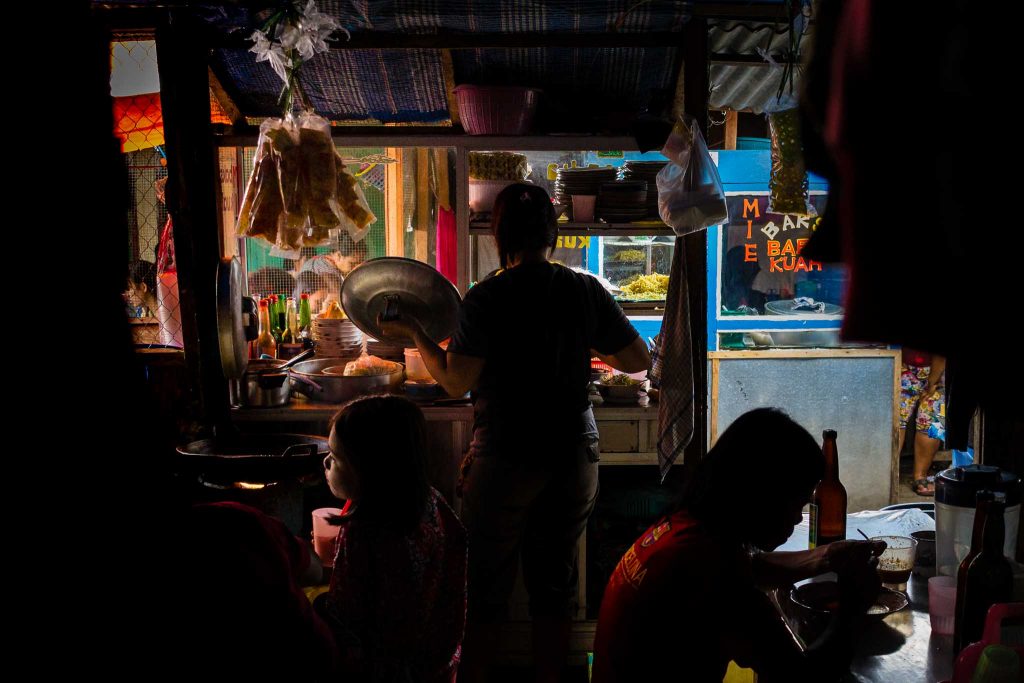 Mercado de comida callejera en Indonesia con gente comiendo en mesas y una mujer abriendo una pota